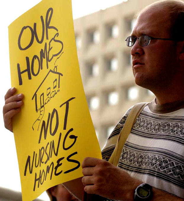 An ADAPT activist holds an Our Homes NOT Nursing Homes sign, at the DUH City Action, September 2008